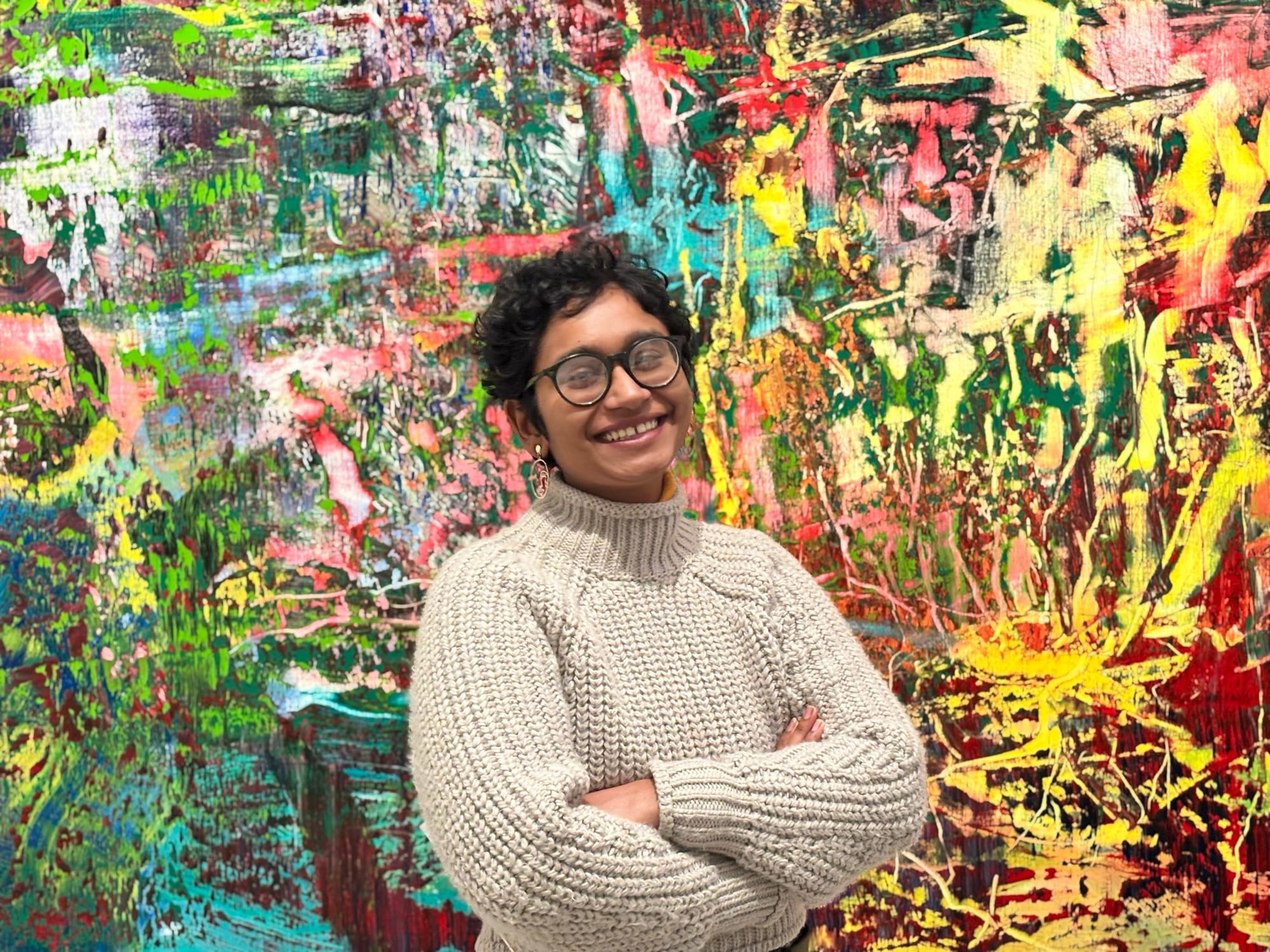 A portrait photo of the author Meera, smiling and standing with crossed arms, infront of a wall painting. She is wearing a black round spectacle and knitted beige sweater.